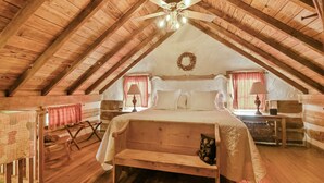 A cozy attic bedroom with a wooden ceiling, a bed with white linens, a bench, and a small table with a lamp.