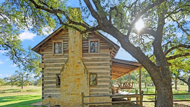 A rustic log cabin with a stone chimney, a covered porch, and a large tree in the foreground.