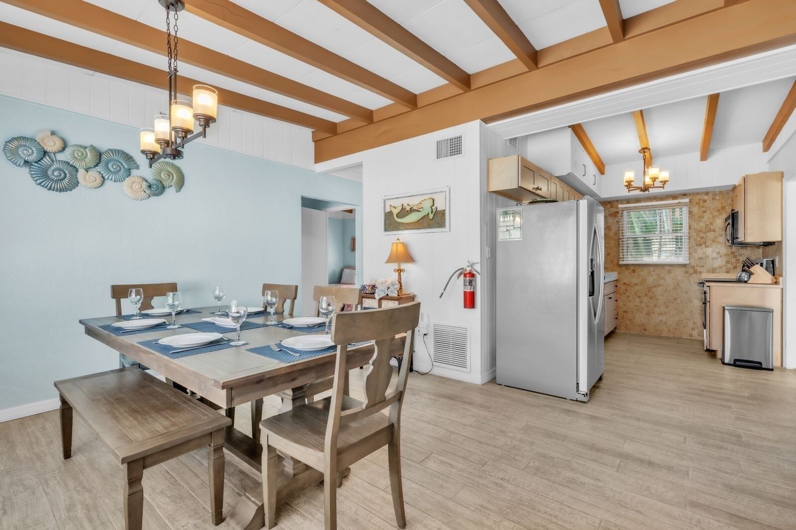 Een eetgedeelte met een houten tafel gedekt voor vier personen, een keuken met een koelkast en kasten, en een plafond met zichtbare balken.