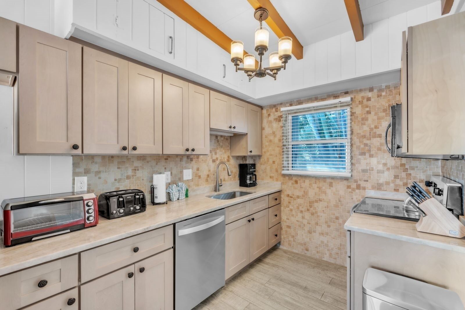 A modern kitchen with light wood cabinets, a built-in dishwasher, a double sink, and a window with blinds.