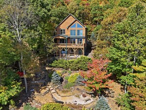 Une maison en bois à deux étages, dotée d’un balcon, entourée d’arbres et bordée d’un petit étang.
