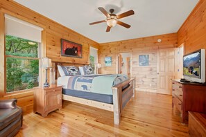 A wooden cabin bedroom with a bed, a ceiling fan, a dresser, and a television.