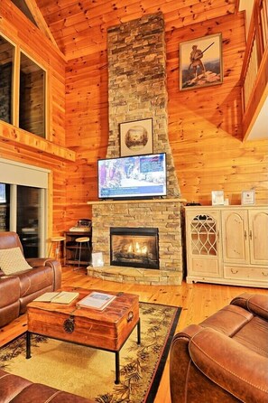 A cozy living room with a stone fireplace, leather furniture, and a television mounted above it. - Sky High in West Jefferson, NC: Spacious 4BR cabin for 10 with stunning mountain views. (West Jefferson)