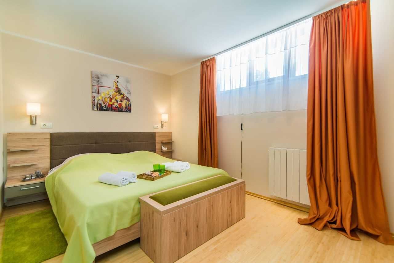 A hotel room with a bed, a wooden bench, a radiator, and a window with curtains.