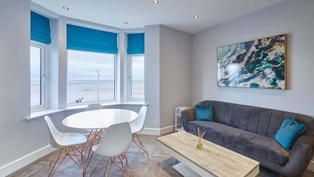 Un salon moderne avec un canapé gris, des chaises blanches, une table basse en bois et un grand tableau abstrait accroché au mur.