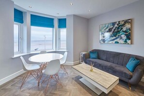 Een moderne woonkamer met een grijze bank, witte stoelen, een houten salontafel en een groot abstract schilderij aan de muur.