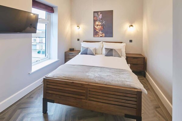 Een slaapkamer met een houten bed, twee nachtkastjes, een wandtelevisie en een raam met uitzicht op gebouwen.