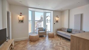 A modern living room with a herringbone floor, a sofa, a chair, two round wicker ottomans, and a television on a wooden stand. - Host & Stay | Abbey Hus (Whitby)