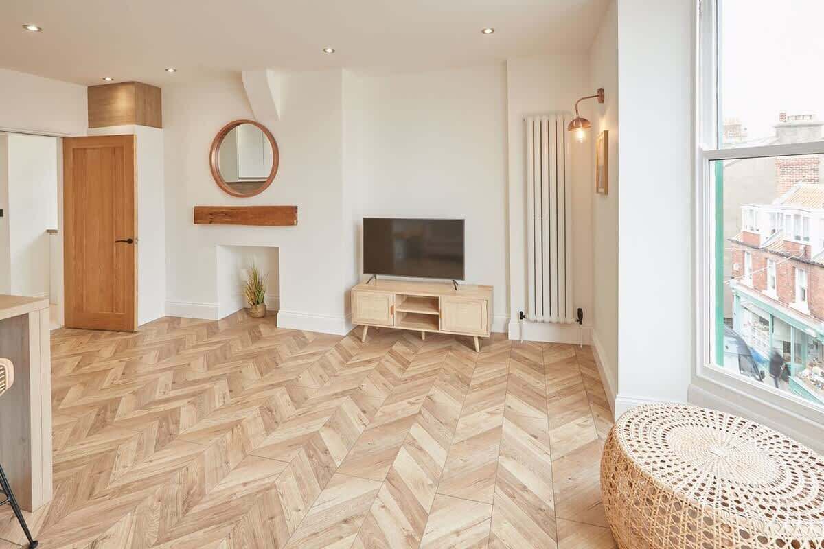 A modern living room with a herringbone wooden floor, a television on a wooden stand, a round mirror on the wall, and a window with a view of buildings.