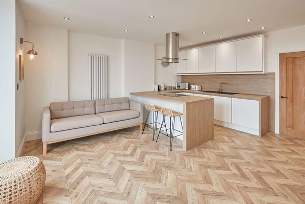 Une cuisine moderne avec un parquet disposé en chevrons, un îlot central doté de tabourets, un canapé et un radiateur.