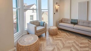 A modern living room with a sofa, armchair, and two woven ottomans, large windows, and a radiator. - Host & Stay | Abbey Hus (Whitby)