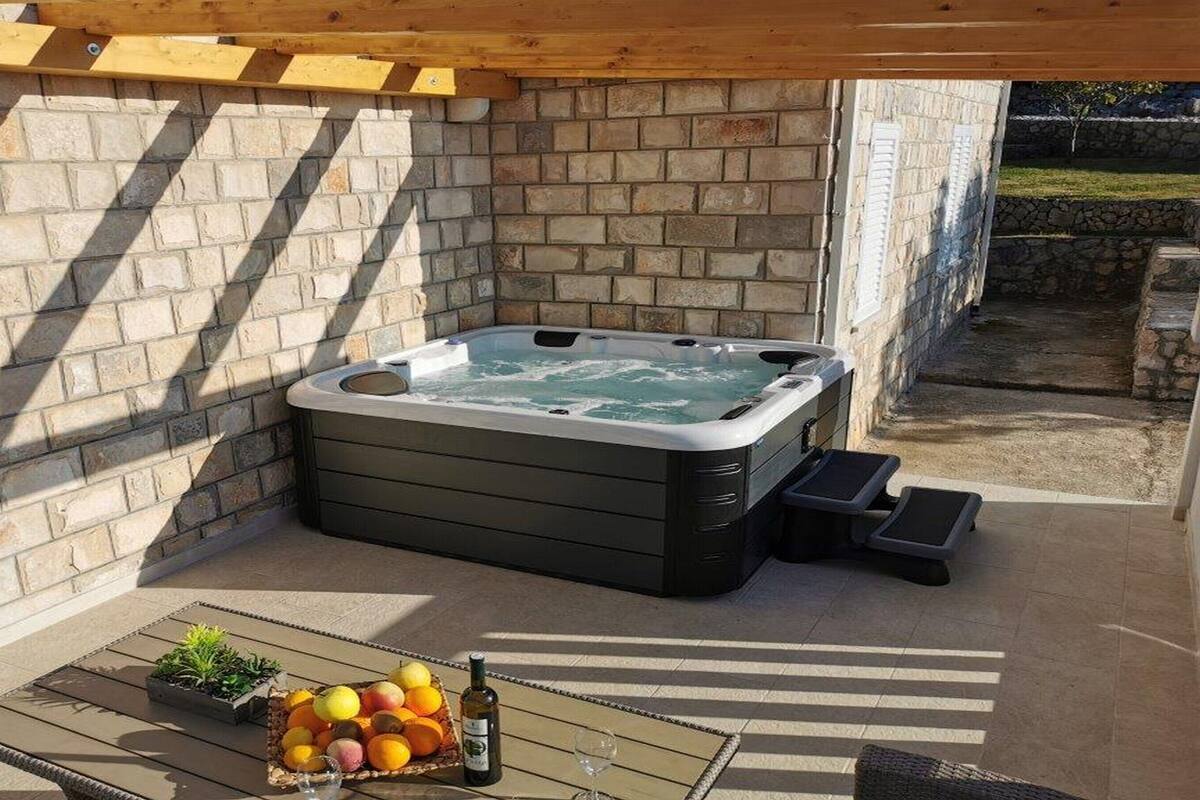 A hot tub in a covered outdoor area with a brick wall, a small table with fruits and a bottle, and a wooden bench.