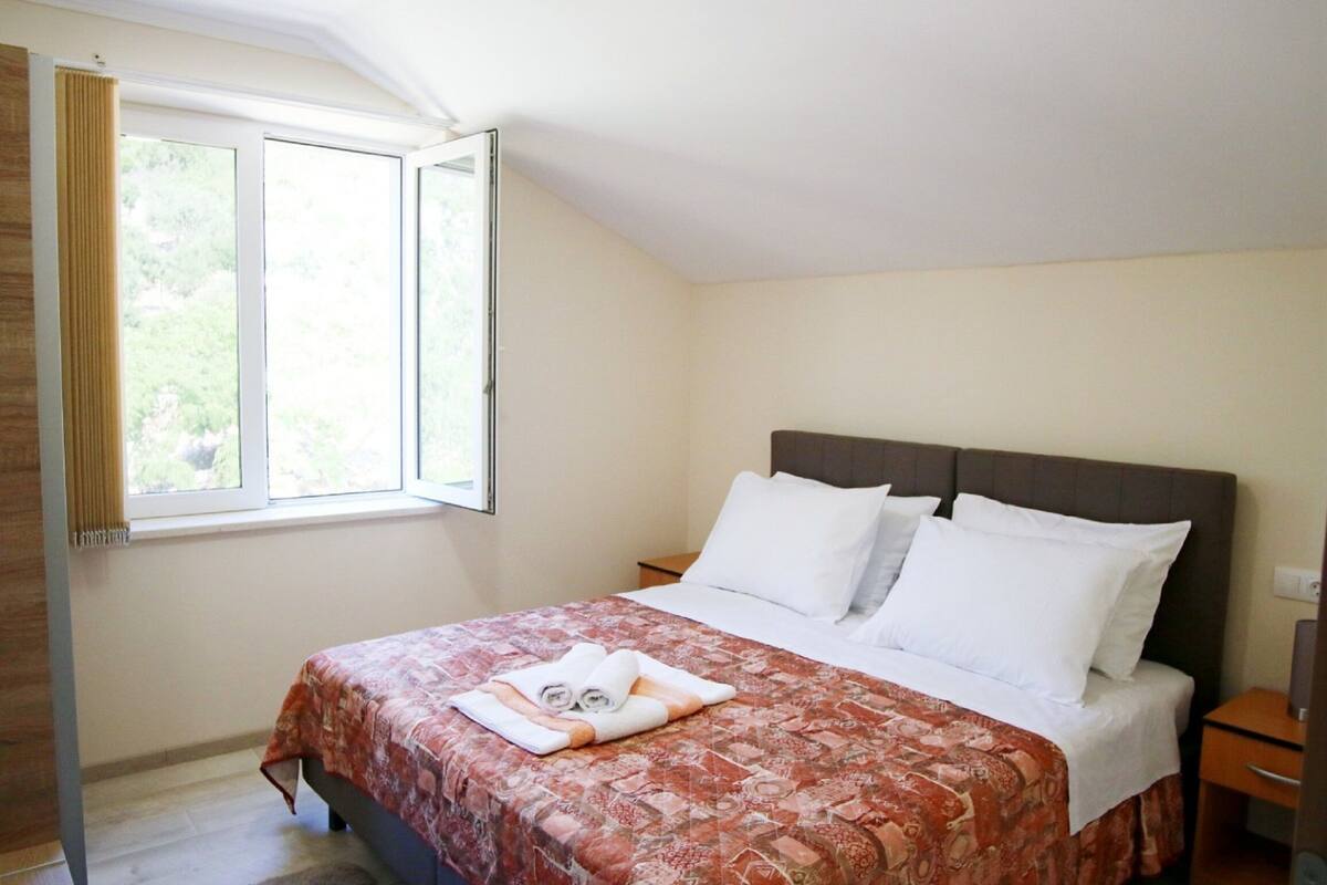 A neatly made bed with a patterned bedspread, a window with curtains, and a nightstand with a lamp.