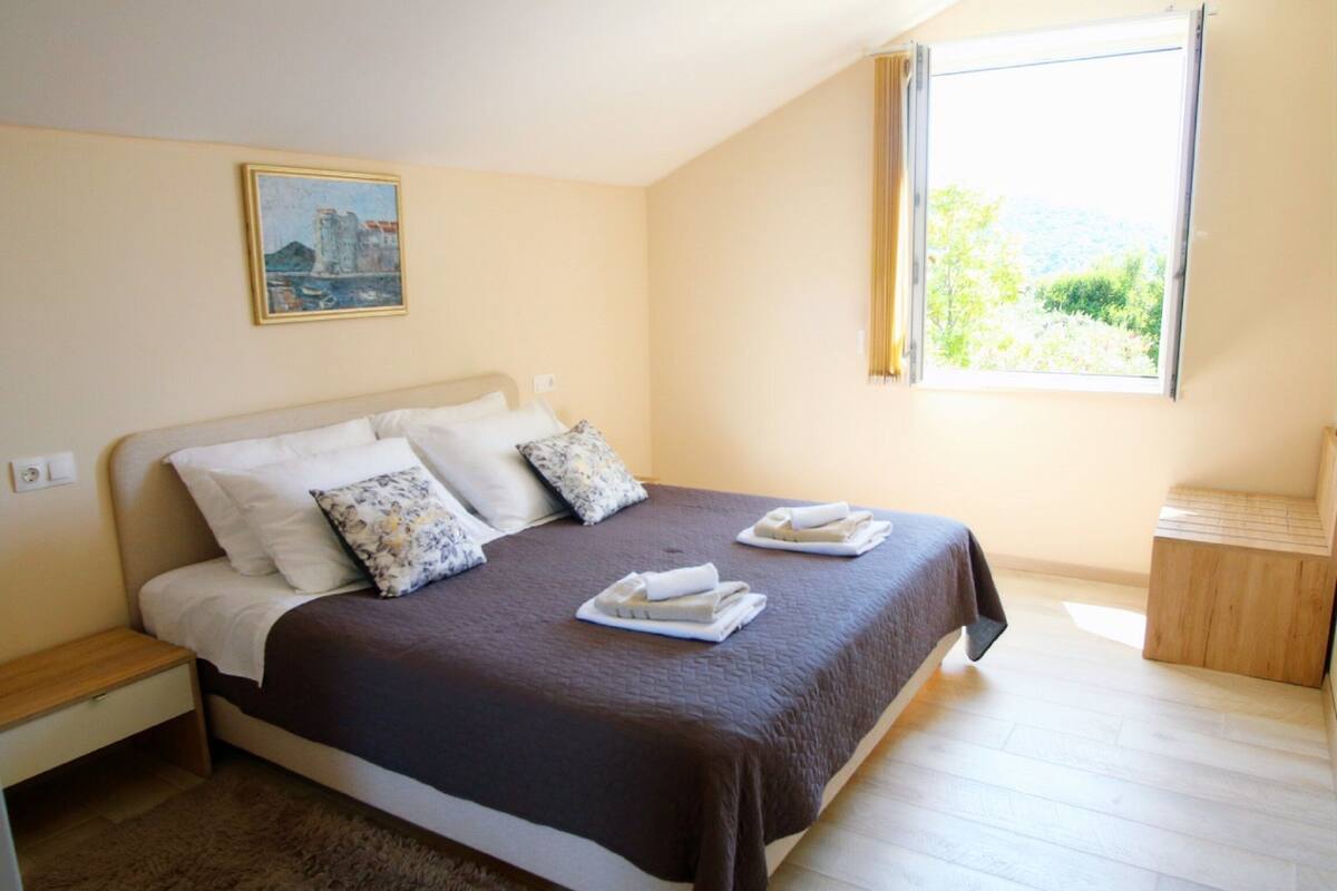 A neatly made bed with a brown bedspread and white pillows, a small bedside table, and a window with a view of greenery.
