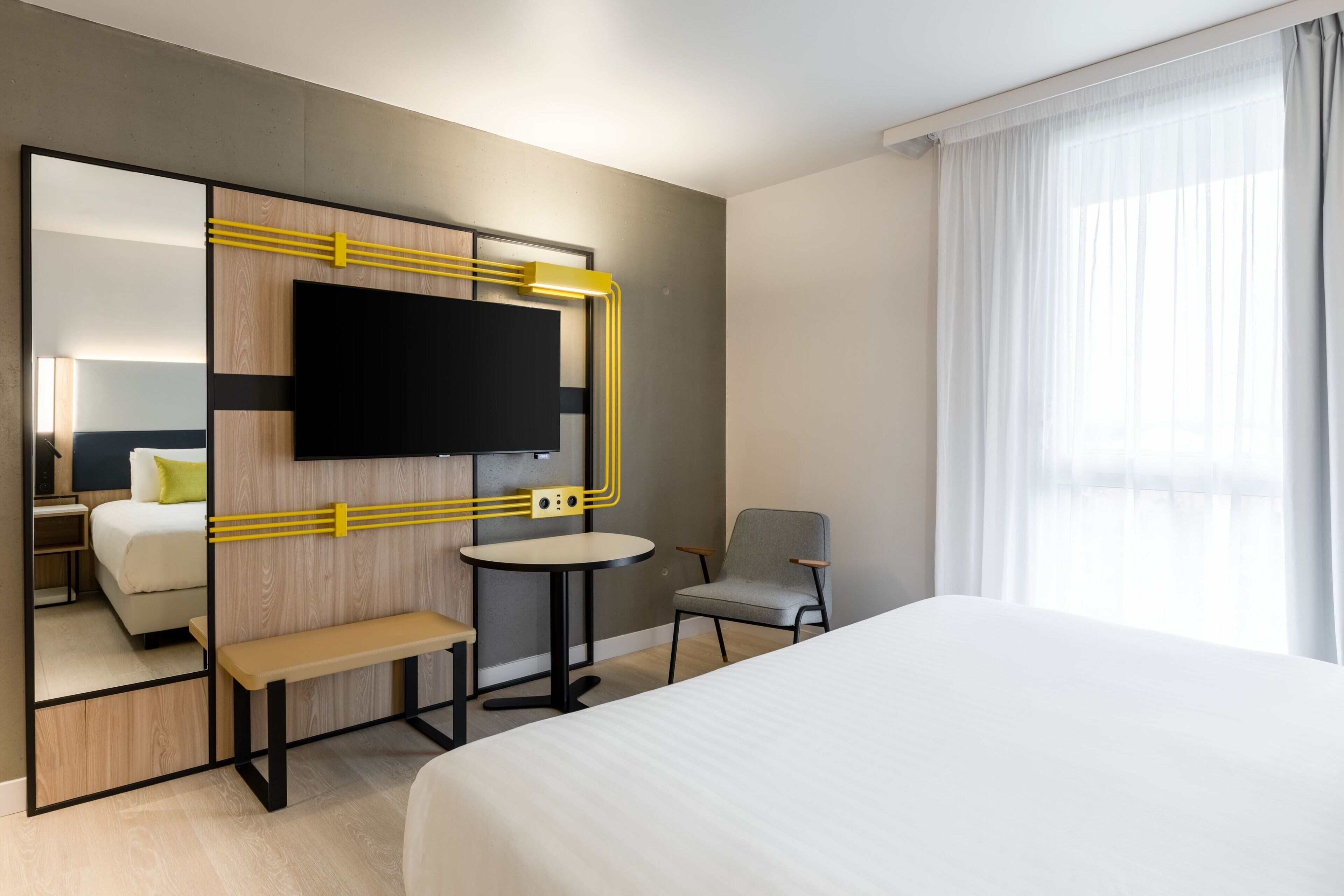 A hotel room with a large bed, a desk with a chair, a television mounted on a wooden panel, and a window with sheer curtains.