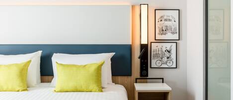 Una cama bien hecha con almohadas blancas y amarillas, un cabecero de madera y una mesita de noche con una lámpara. Hay dos cuadros en la pared.