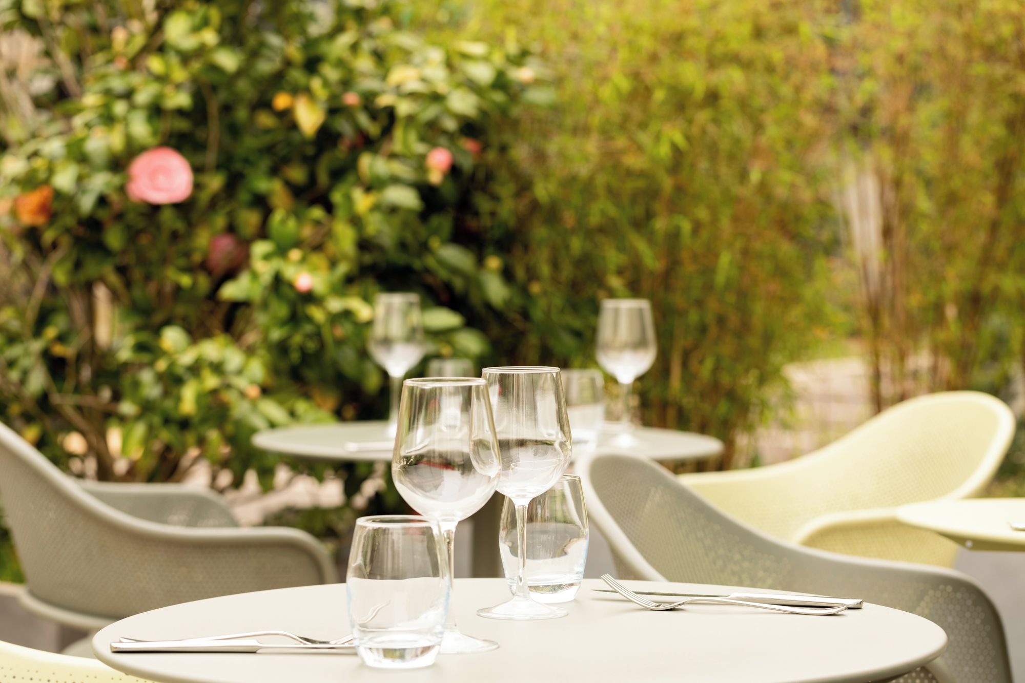 Une table dressée avec des verres à vin et des couverts, entourée de chaises, dans un cadre extérieur avec de la verdure en arrière-plan.