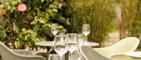 Une table dressée avec des verres à vin et des couverts, entourée de chaises, dans un cadre extérieur avec de la verdure en arrière-plan.
