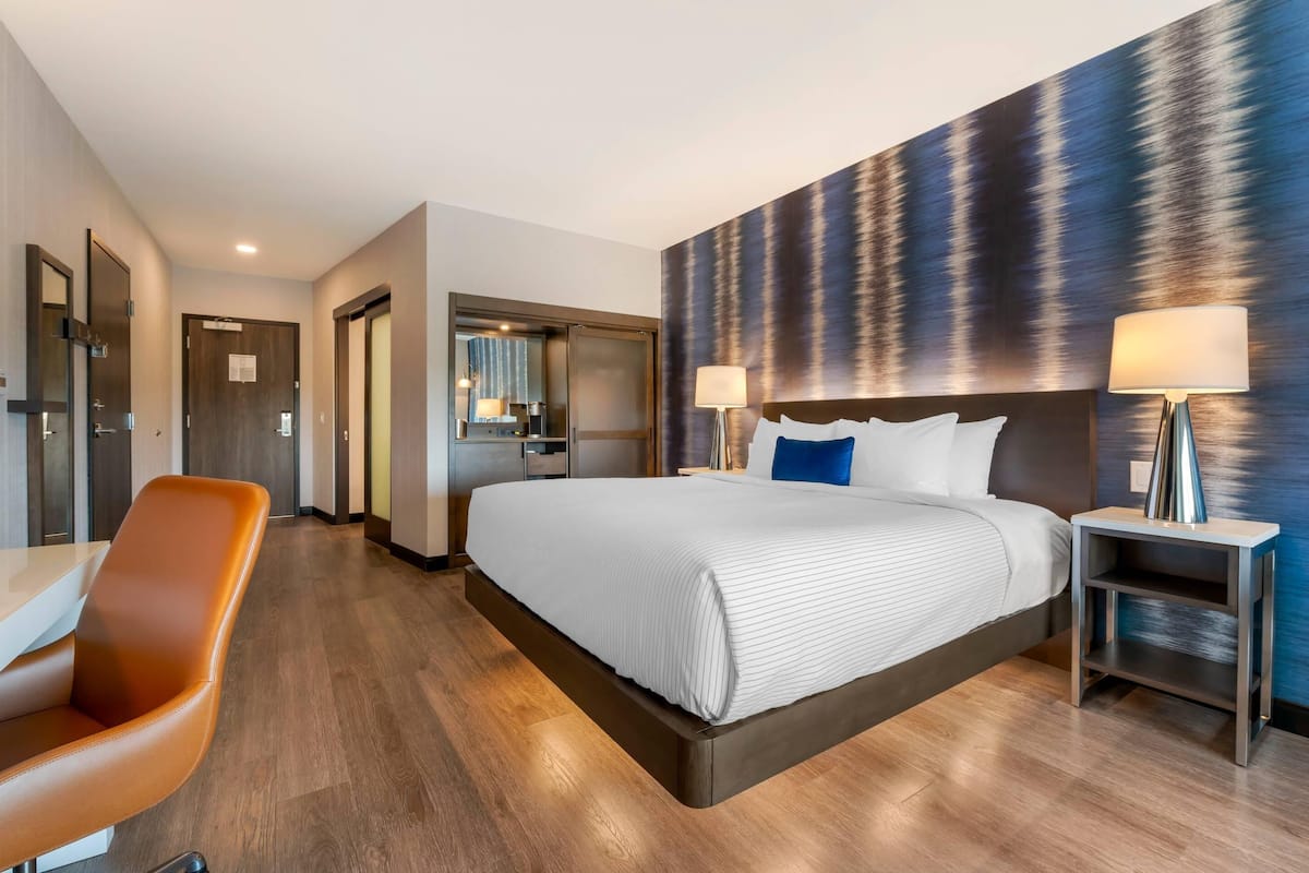 Una habitación de hotel moderna con una cama grande, un escritorio y una silla. Cuenta con un baño con espejo y ducha.
