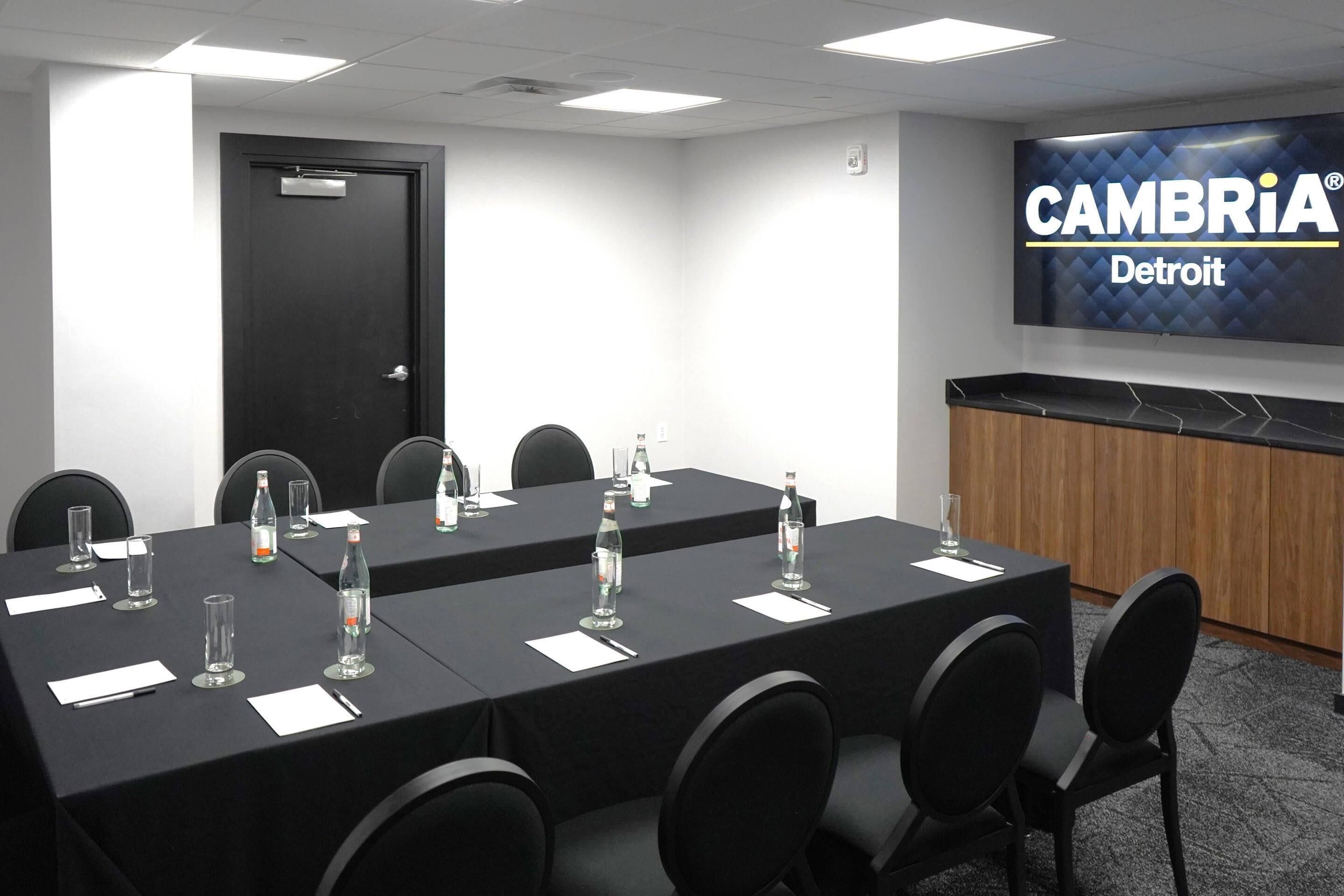 A conference room with a long table, chairs, water bottles, and a presentation screen displaying "CAMBRIA Detroit".