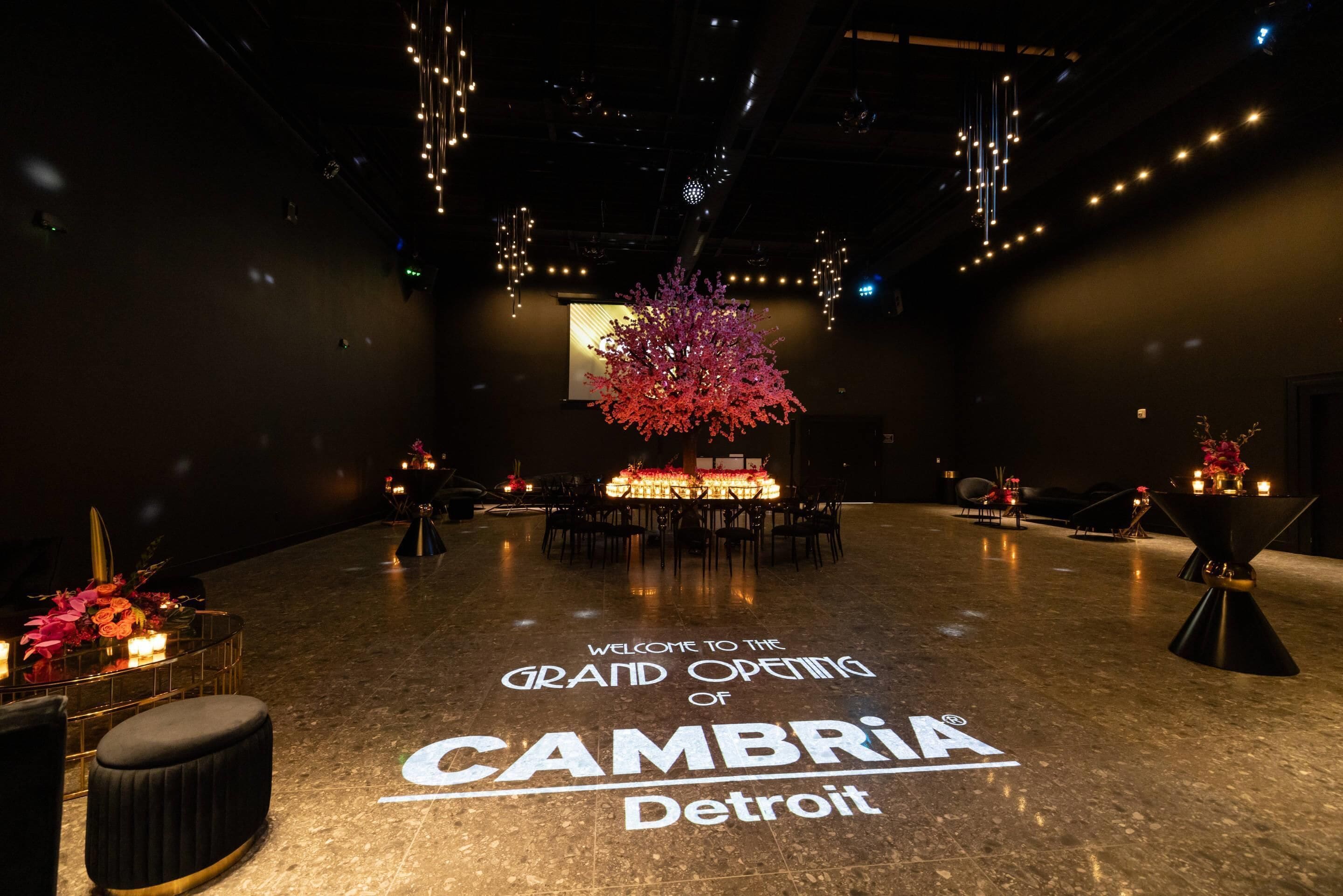 A spacious hall with a welcoming message for the grand opening of CAMBRIA in Detroit, featuring a central floral arrangement and tables set for an event.