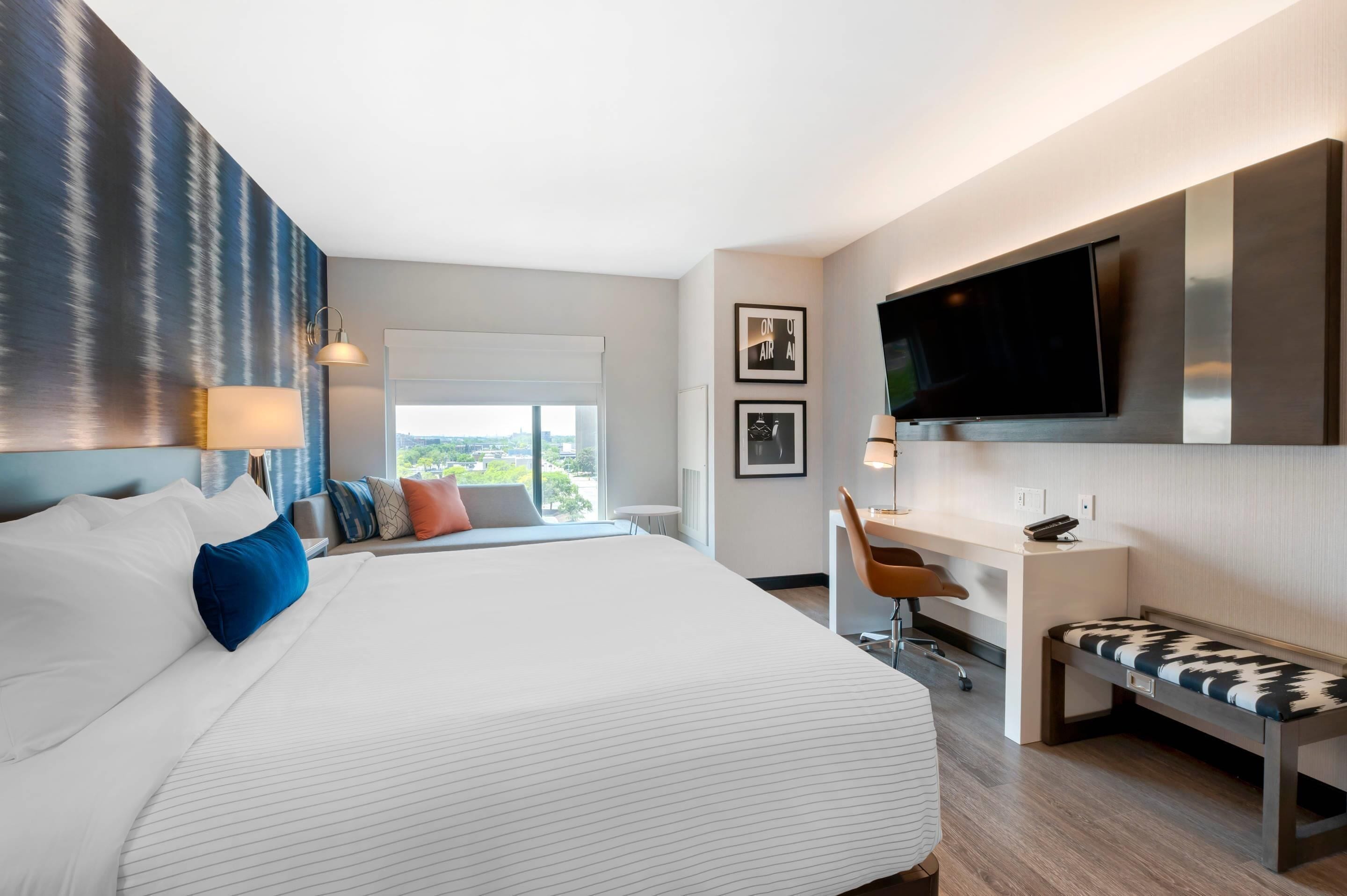 Habitación de hotel moderna con una cama grande, un escritorio, un banco, un televisor y una ventana con vistas a la ciudad.
