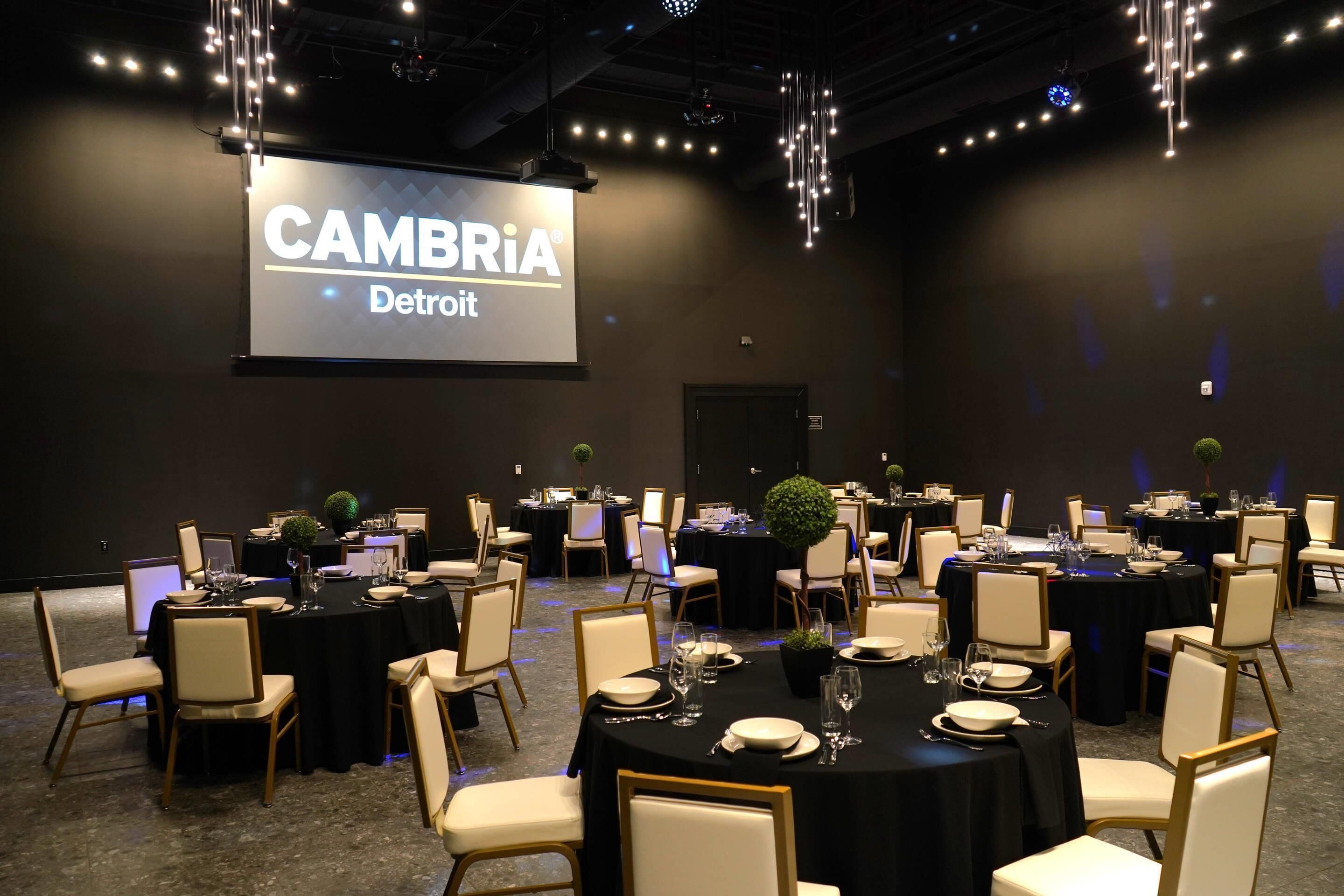 A conference room with multiple round tables, each set with white chairs and black tablecloths. A large screen displays "CAMBRIA Detroit".