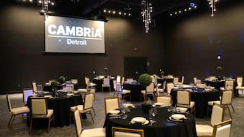 Sala de conferência com várias mesas redondas, cada uma com cadeiras brancas e toalhas de mesa pretas. Uma tela grande exibe "CAMBRIA Detroit".