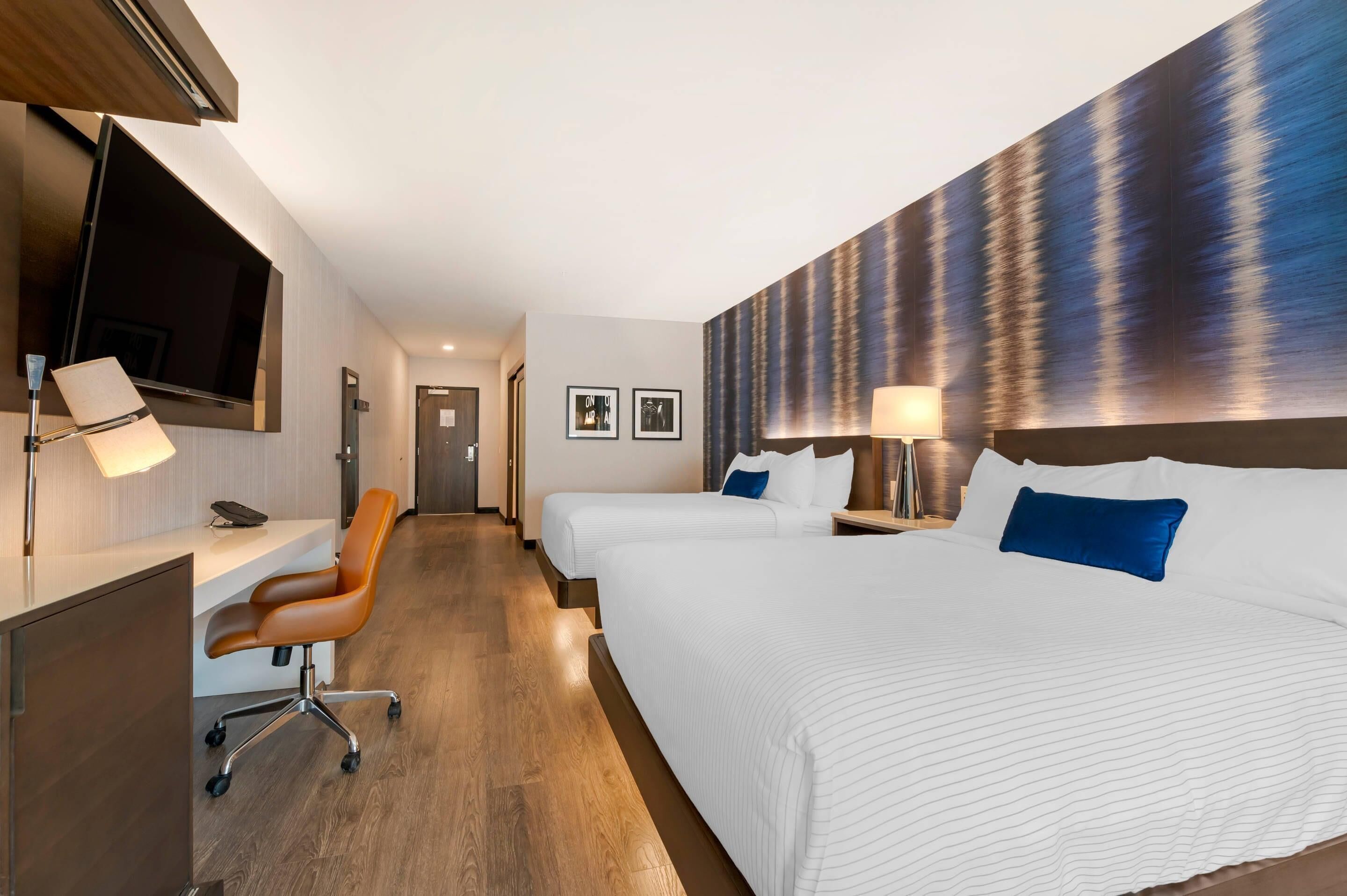 A hotel room with a large bed, a desk with a chair, a TV mounted on the wall, and a painting on the wall.