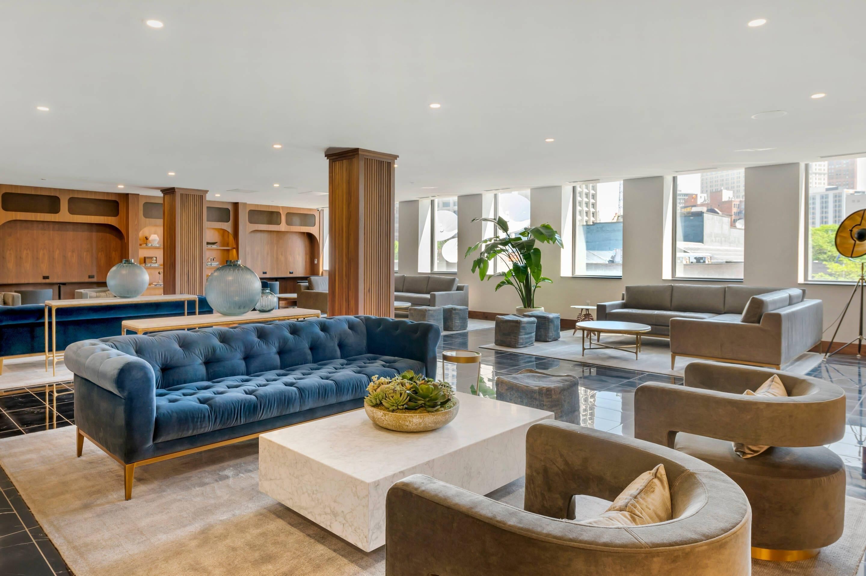 A spacious lobby with a blue sofa, grey armchairs, and a coffee table. There are large windows providing natural light and a view of the city.