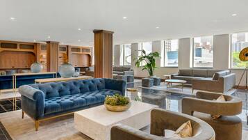 A spacious lobby with a blue sofa, grey armchairs, and a coffee table. There are large windows providing natural light and a view of the city.