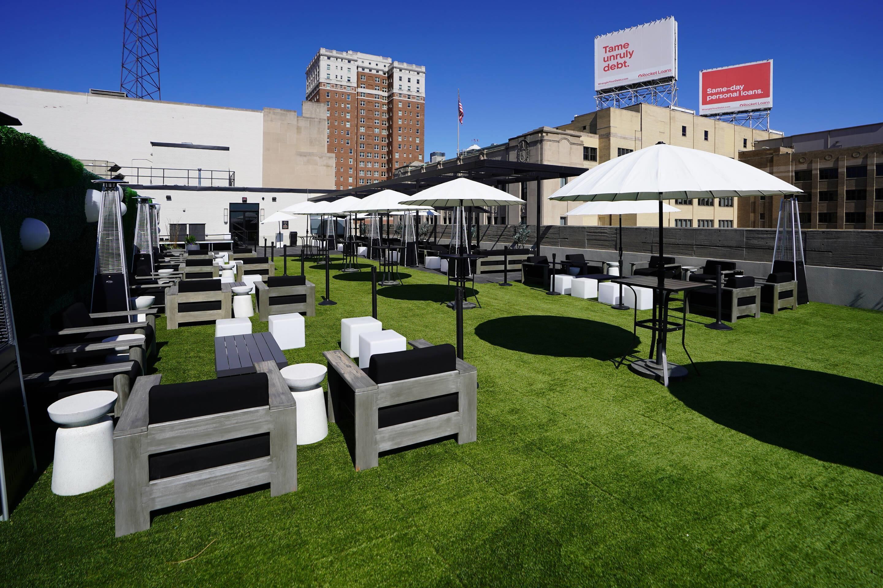 Een buitenruimte met tafels en stoelen, omringd door kunstgras, onder parasols, met een stadsgezicht op de achtergrond.