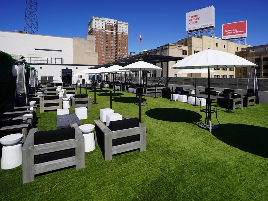 Een buitenruimte met tafels en stoelen, omringd door kunstgras, onder parasols, met een stadsgezicht op de achtergrond.
