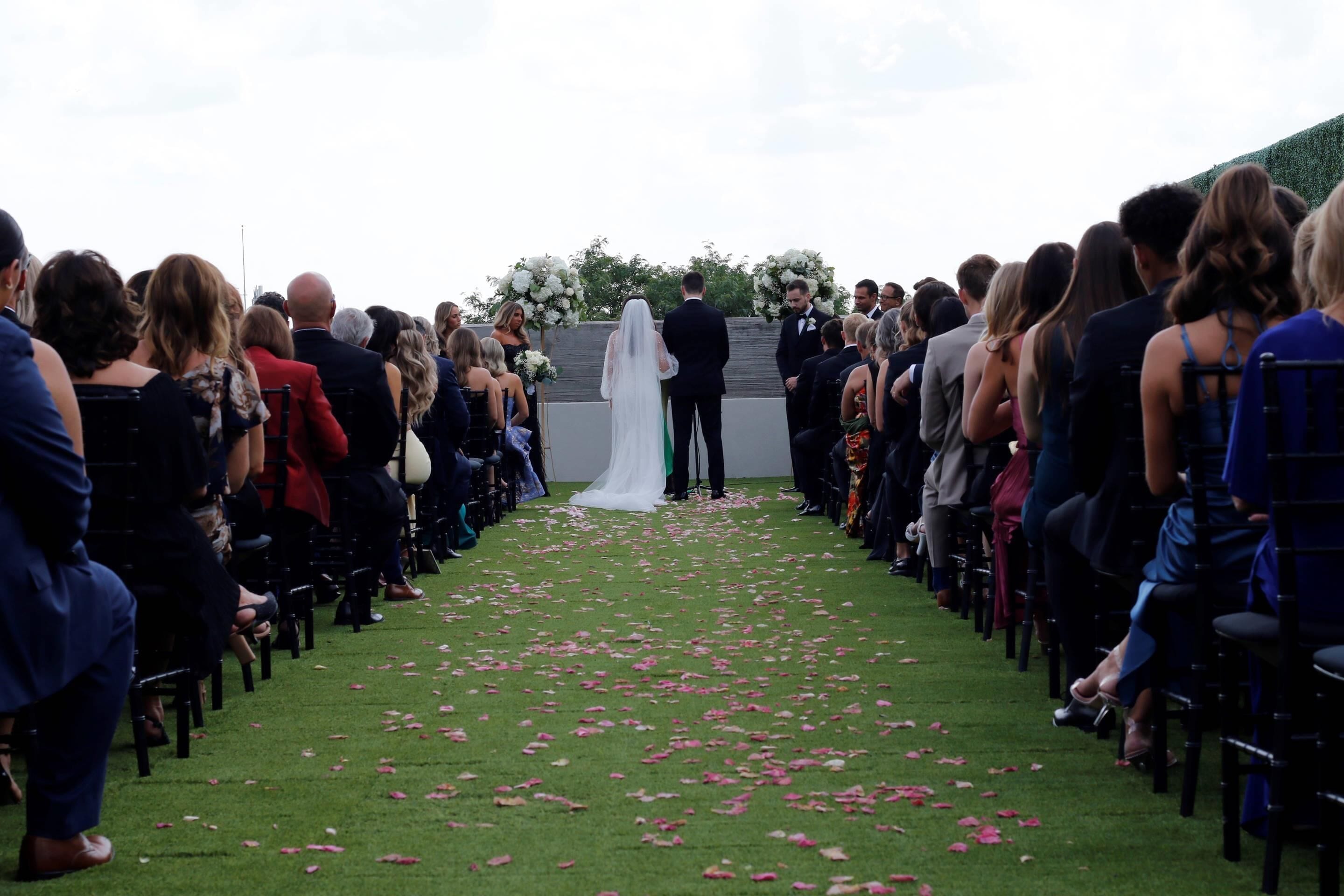 Una ceremonia de boda con invitados sentados en sillas frente a un pasillo verde adornado con pétalos de rosa.