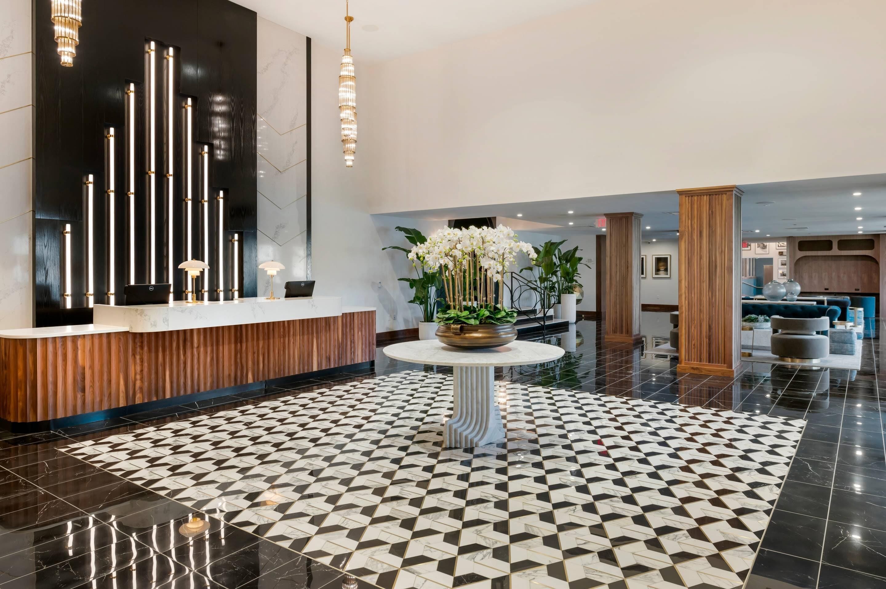 A modern hotel lobby with a black and white tiled floor, a reception desk, a round table with a vase of white flowers, and a chandelier.