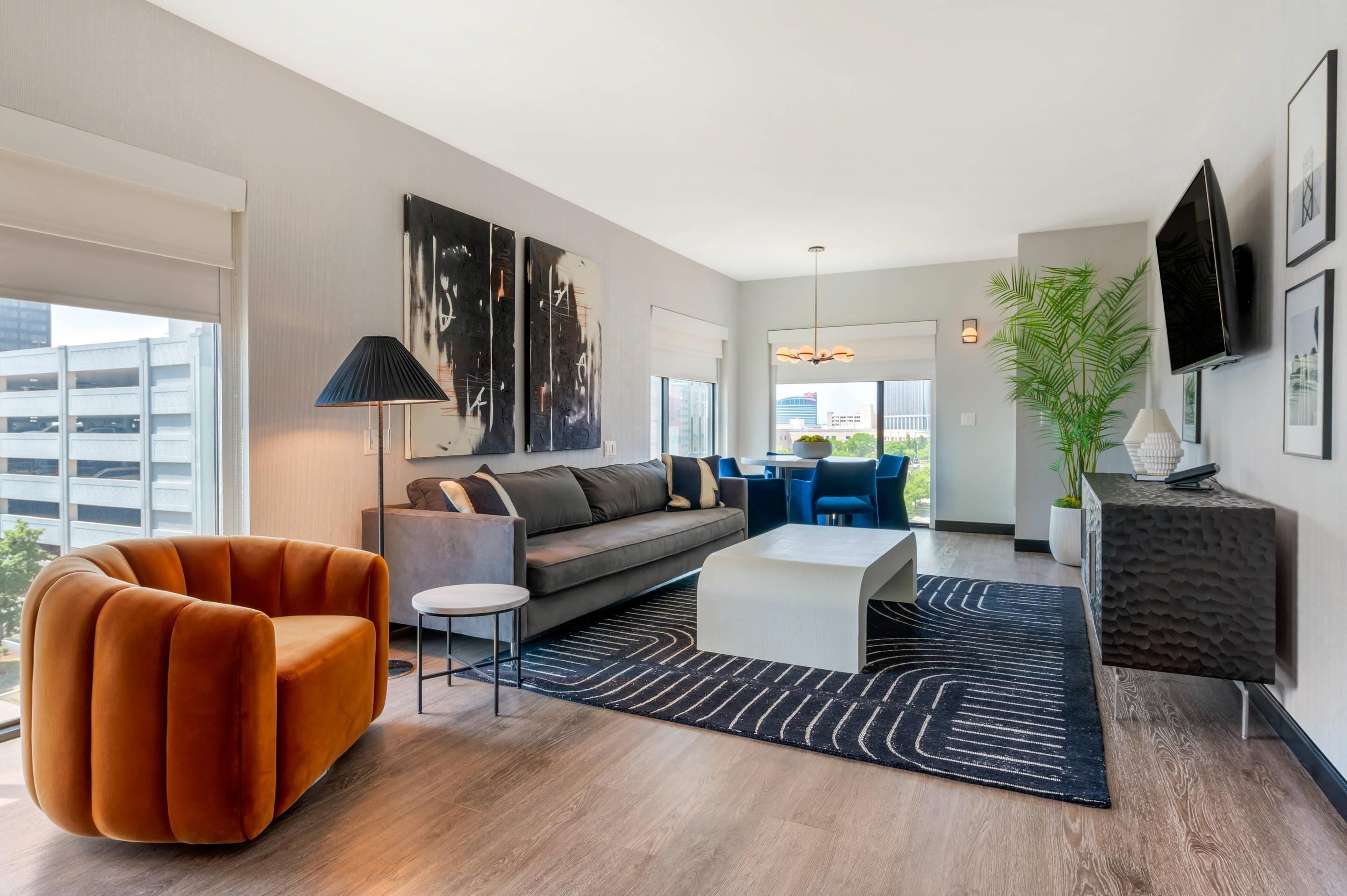 A modern living room with a sofa, orange armchair, and a coffee table. There is a large window, a plant, and a TV mounted on the wall.