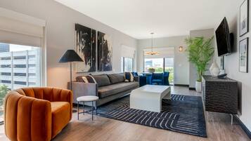 A modern living room with a sofa, orange armchair, and a coffee table. There is a large window, a plant, and a TV mounted on the wall.