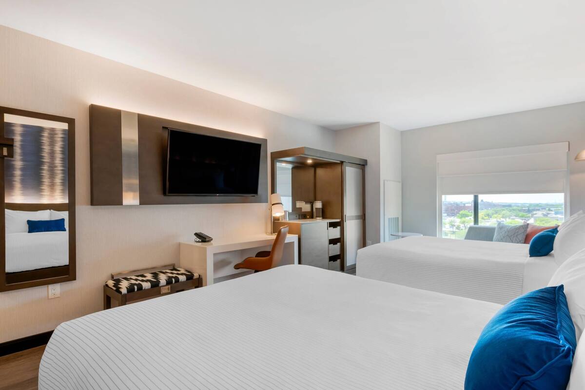 Habitación de hotel con dos camas, un escritorio, televisor, espejo y vistas a la ciudad.