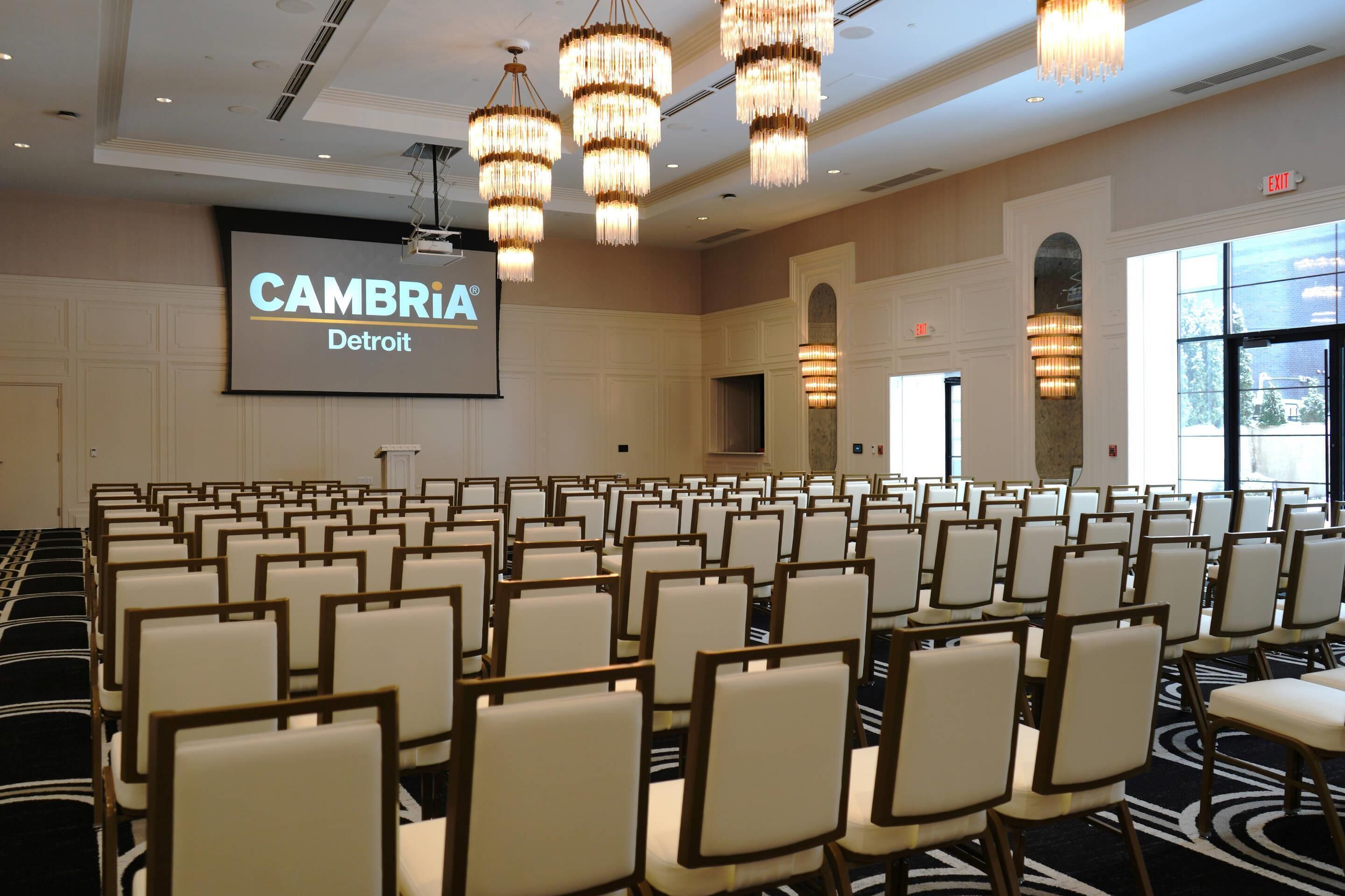 A conference room with rows of chairs, a large screen displaying 'CAMBRIA Detroit', and chandeliers.