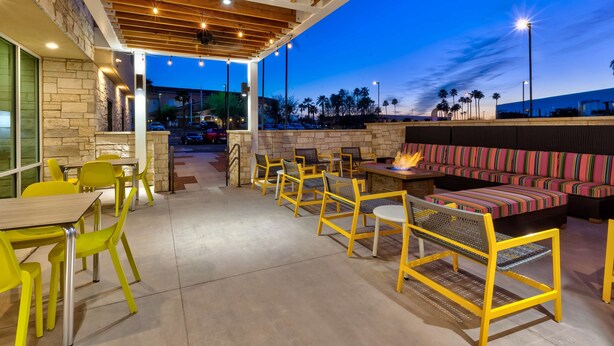 An outdoor seating area with yellow chairs and tables, a fire pit, and a stone wall. - Home2 Suites by Hilton Tucson Airport (Tucson)