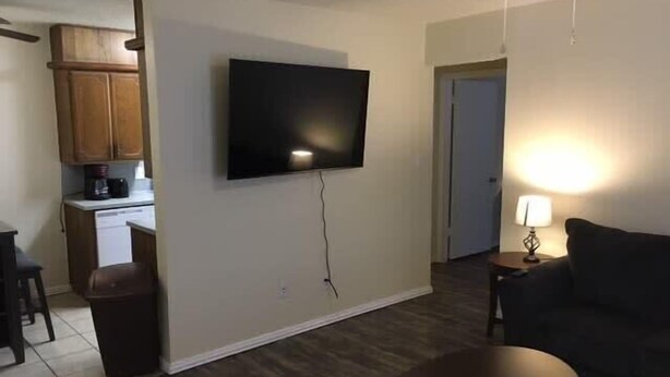 Een woonkamer met een flatscreen televisie aan de muur, een donkergrijze bank, een klein rond bijzettafeltje en een lamp op een nachtkastje.