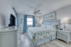 Un dormitorio con cama, ventilador de techo, mesita de noche, cómoda, televisor y ventana con cortinas.
