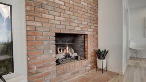 A brick fireplace with a visible fire, a potted plant, and a white chair in a modern room with wooden flooring. - Light + Bright Boulder Midcentury 30+ day rental  (Boulder)