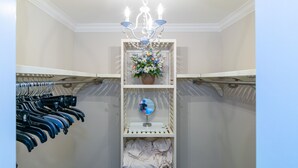Ein begehbarer Kleiderschrank mit Kleidung links, einem Spiegel in der Mitte und einer Chandelierleuchte darüber.