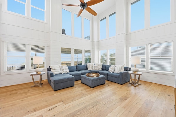 A spacious living room with a sectional sofa, a ceiling fan, large windows, and wooden flooring.