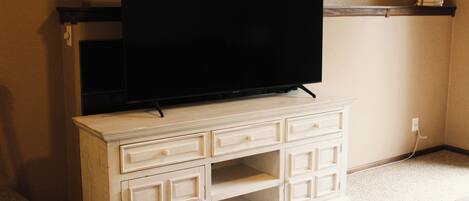A white entertainment center with a flat-screen TV, a shelf above it, and a power outlet on the wall.