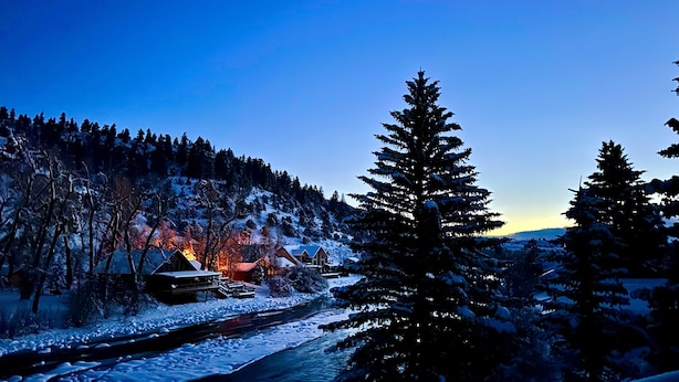 Paisatge cobert de neu amb un riu, un bosc i un edifici amb llums.