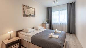 A neatly arranged hotel room with a bed, bedside tables, a nightstand, a lamp, a framed artwork, a dresser, and a window with curtains. - Ferienwohnung am Meer, Urlaub auf der Insel Usedom, Appartement Flora (Swinoujscie)