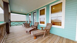A covered porch with wooden chairs and tables, a wooden railing, and a view of a building in the background. - Ocean Jewel: 3  BR, 2  BA House in Port Bolivar, Sleeps 10 (Bolivar Peninsula)
