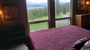 A bedroom with a large window, a bed with a patterned bedspread, a dresser, a lamp, and a framed picture on the wall. - Montana Mountaintop Ranch Estate (Proctor)